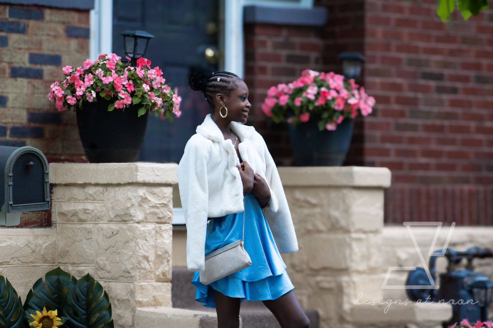 Girl in a white faux fur jacket and blue dress walking past pink flowers on a front stoop