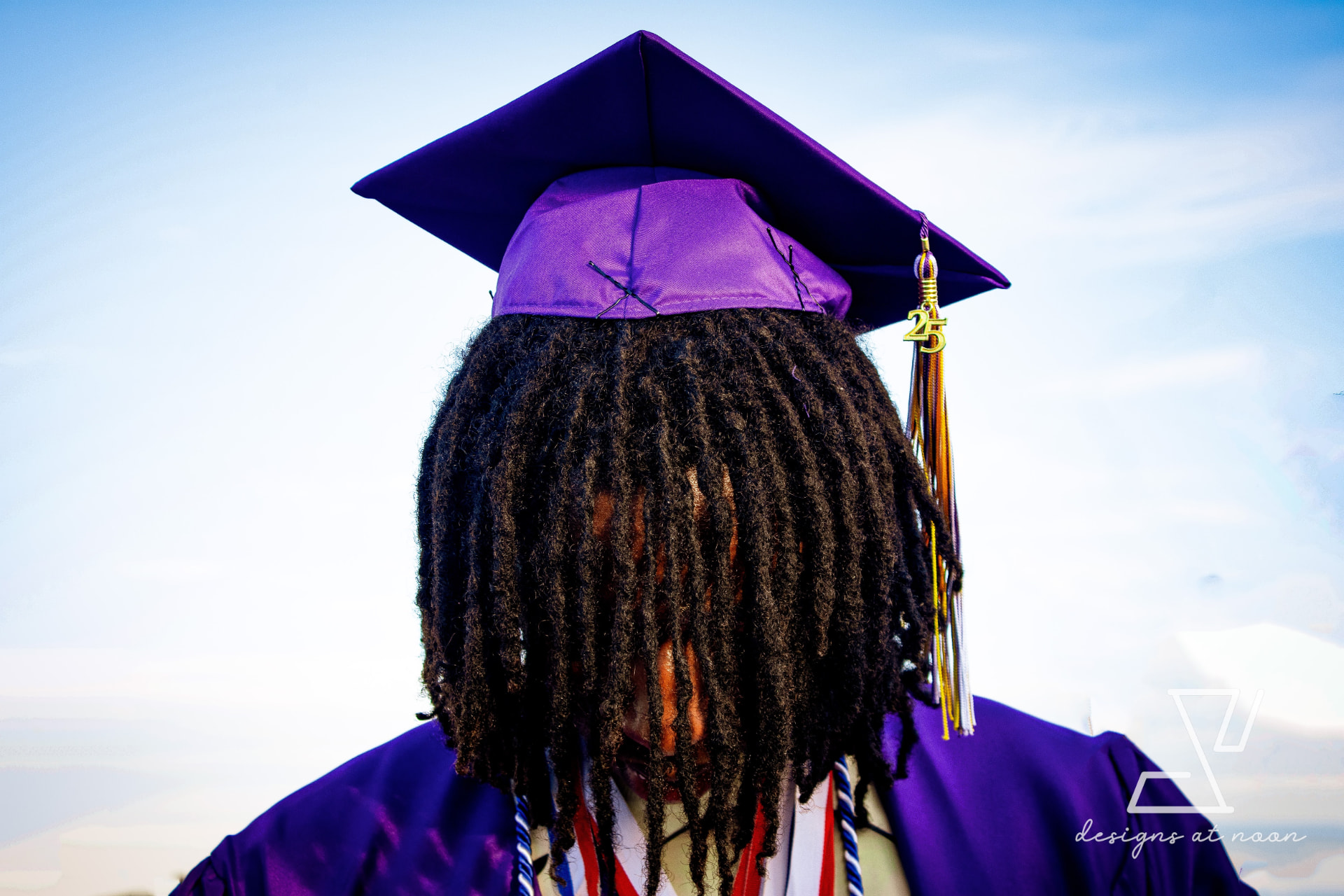 Close portrait of a graduate in a purple cap with a 25 tassel against blue sky