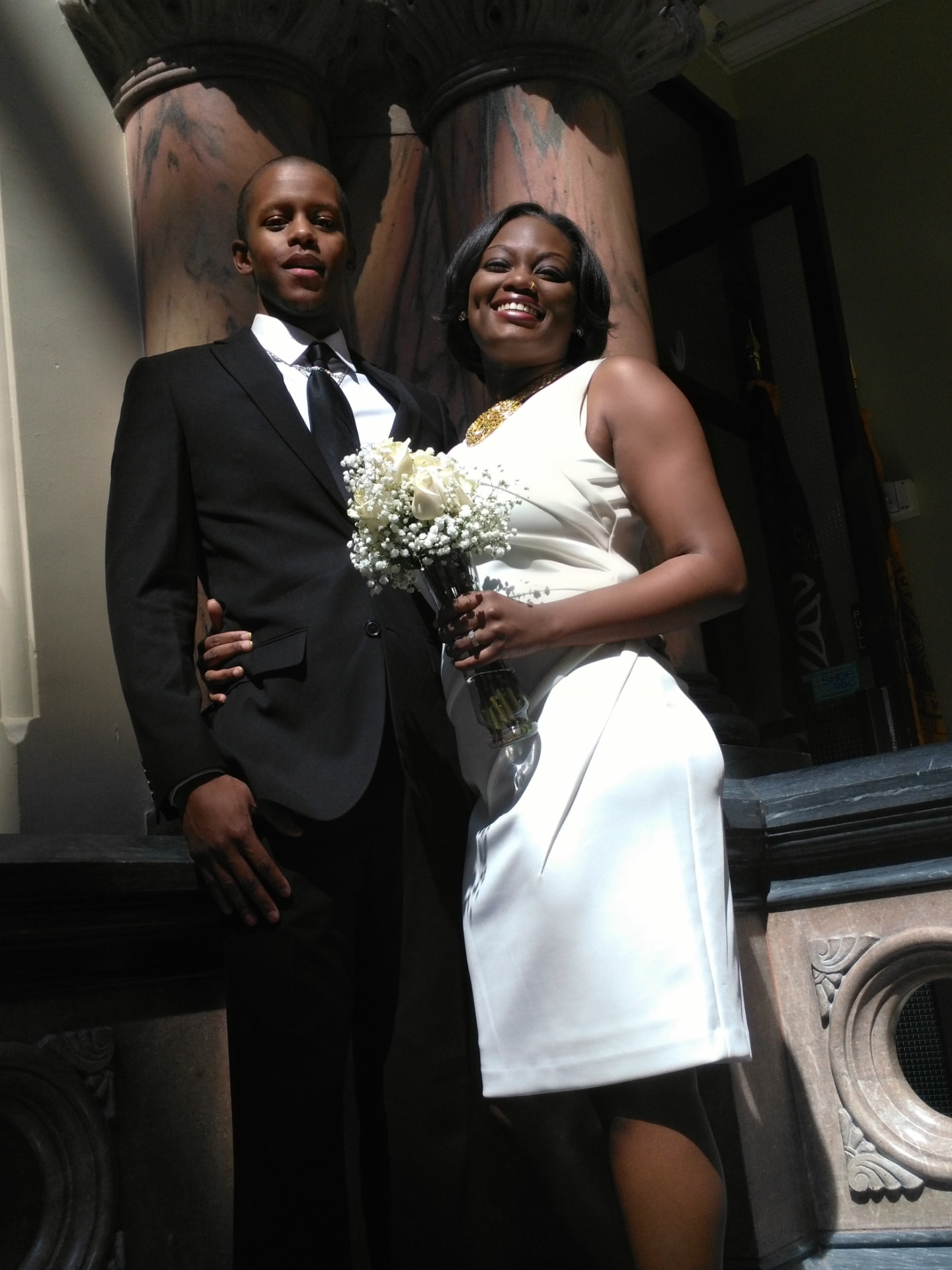Newlywed couple posing with a white rose bouquet beside marble columns