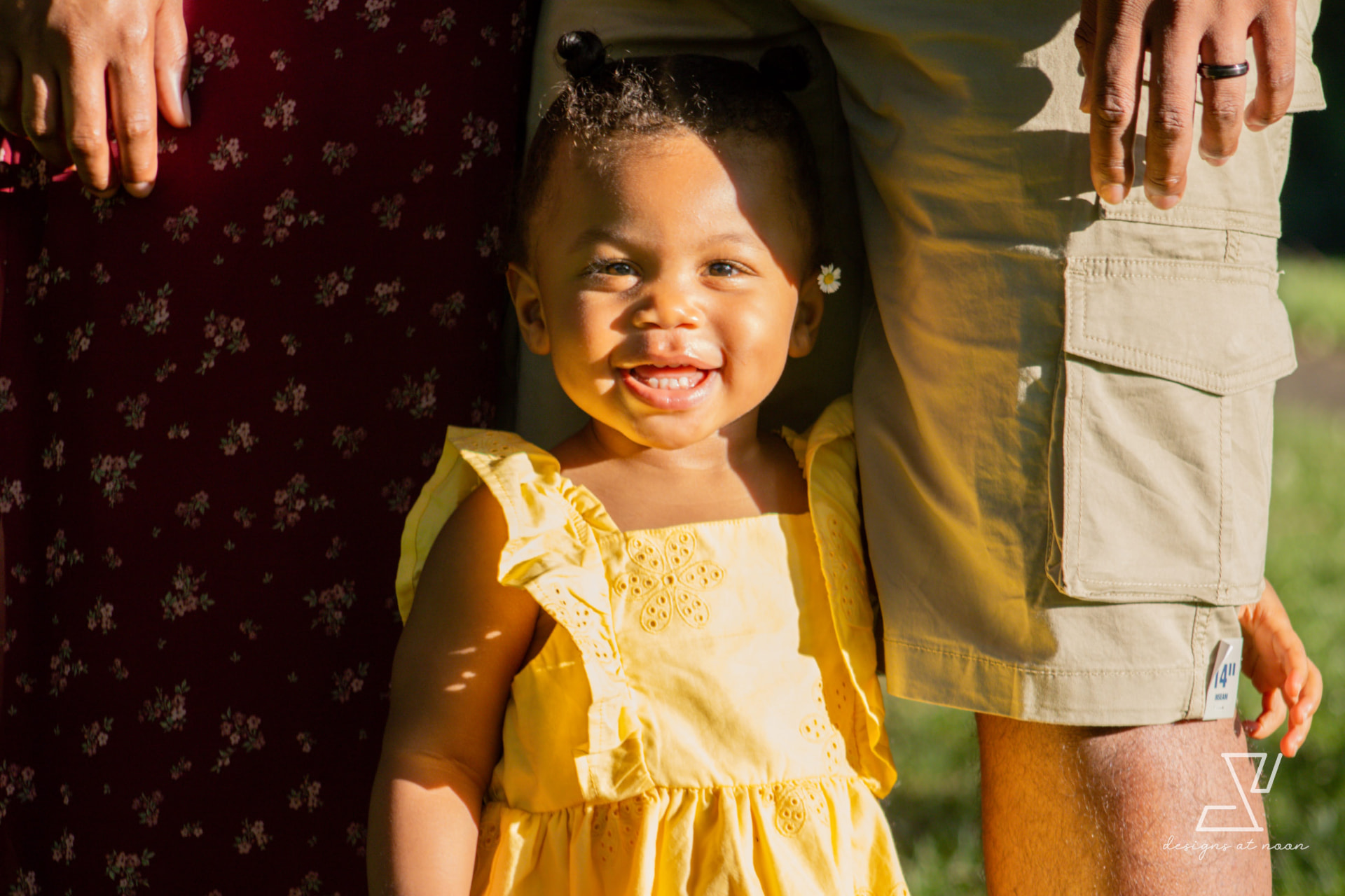 Toddler in a yellow dress smiling between her parents at golden hour
