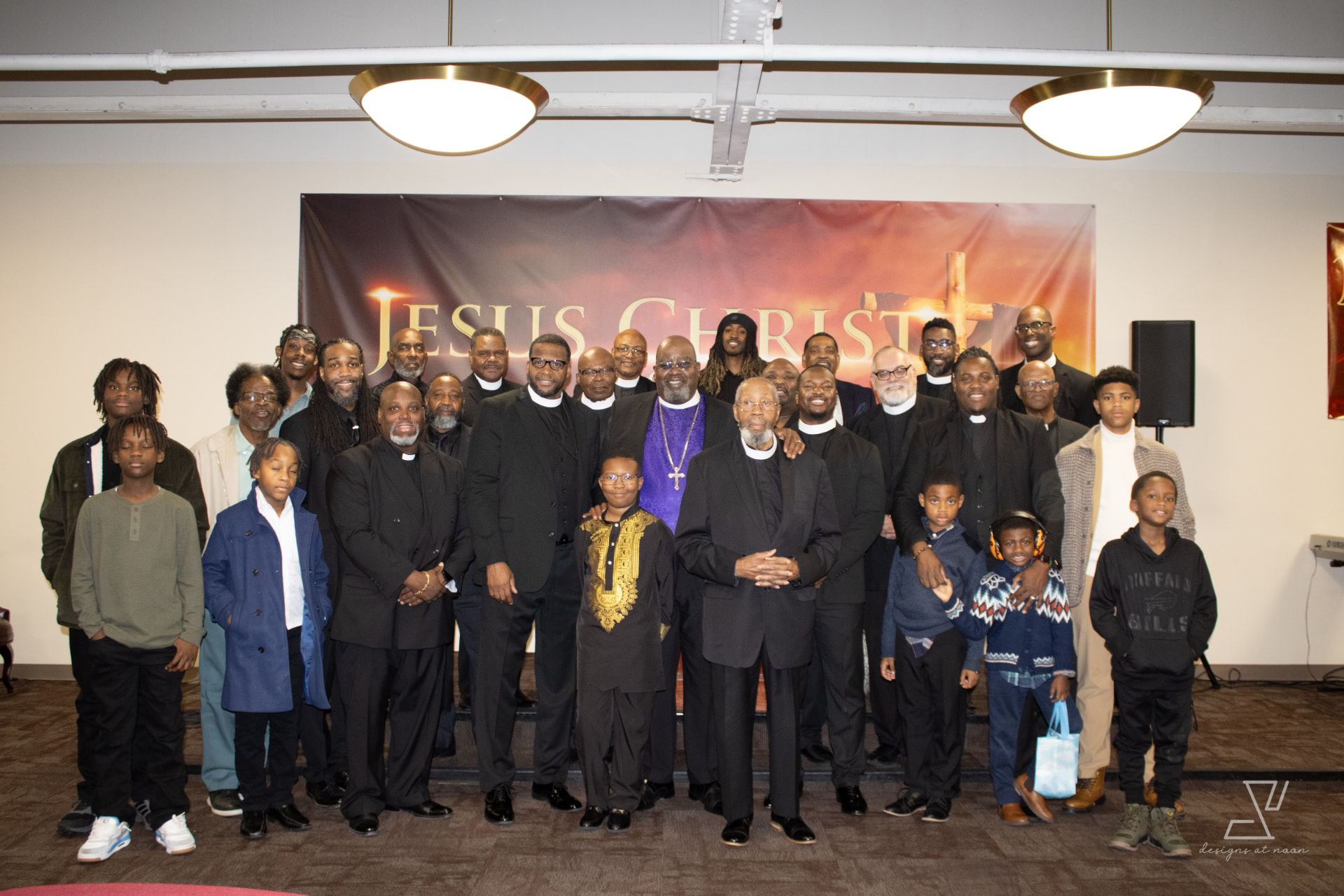 Congregation men and boys gathered for a group portrait in front of a Jesus Christ banner