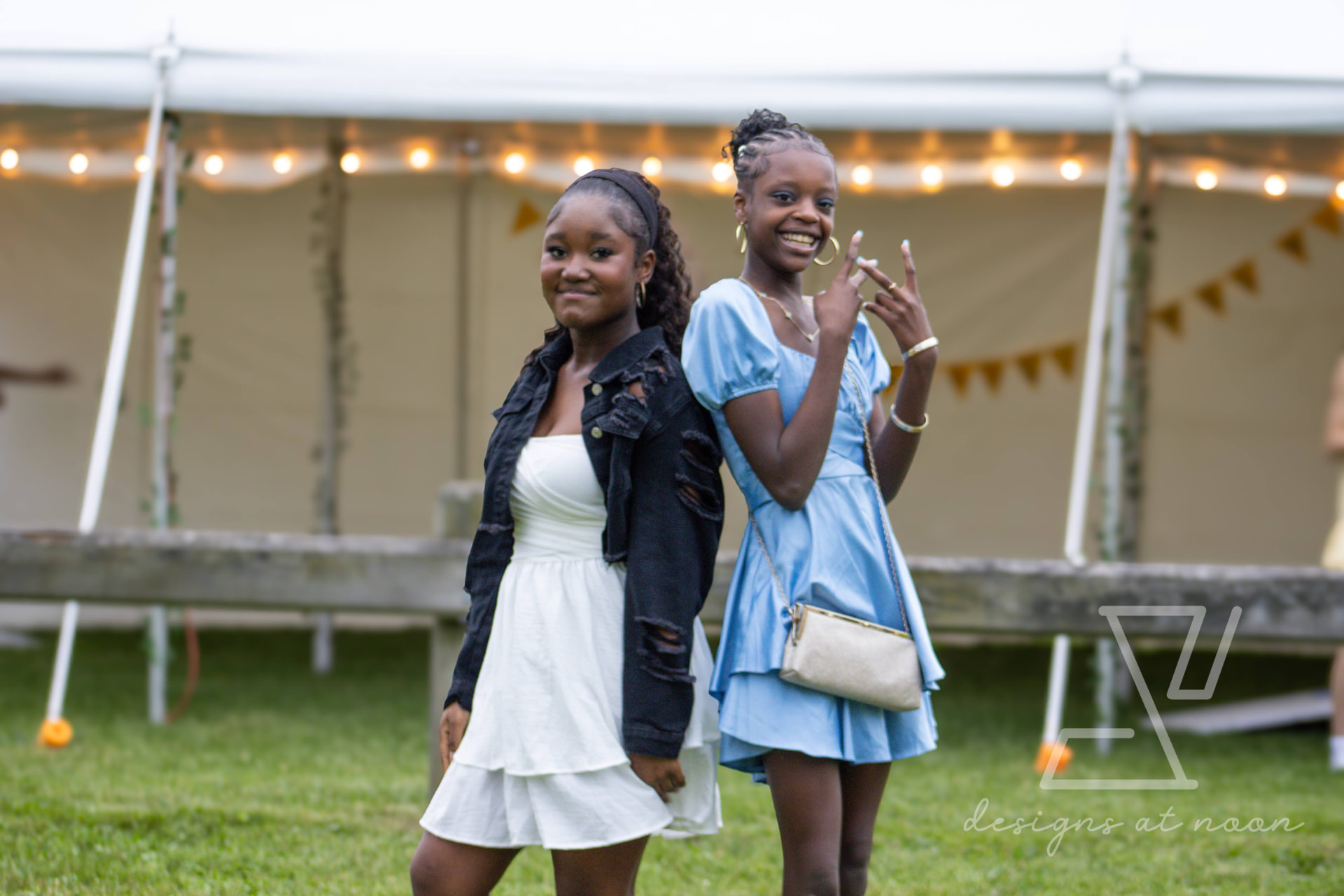 Two girls posing back to back under string lights at a backyard tent celebration